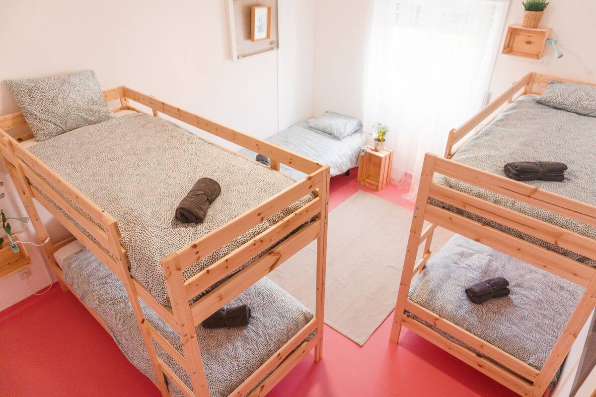 Een kamer met drie stapelbedden, een klein nachtkastje met een plantje, en een raam met gordijnen.