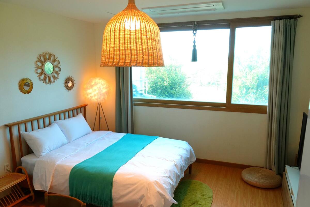 A bedroom with a bed, a standing lamp, a window with curtains, and a view of greenery outside.