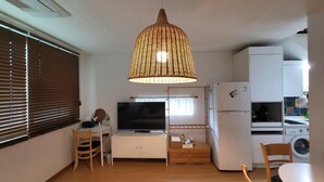 A compact living space with a TV, a small kitchenette, a dining area, and a large woven light fixture. - Jeju Eltzutown Pension (Jeju City)
