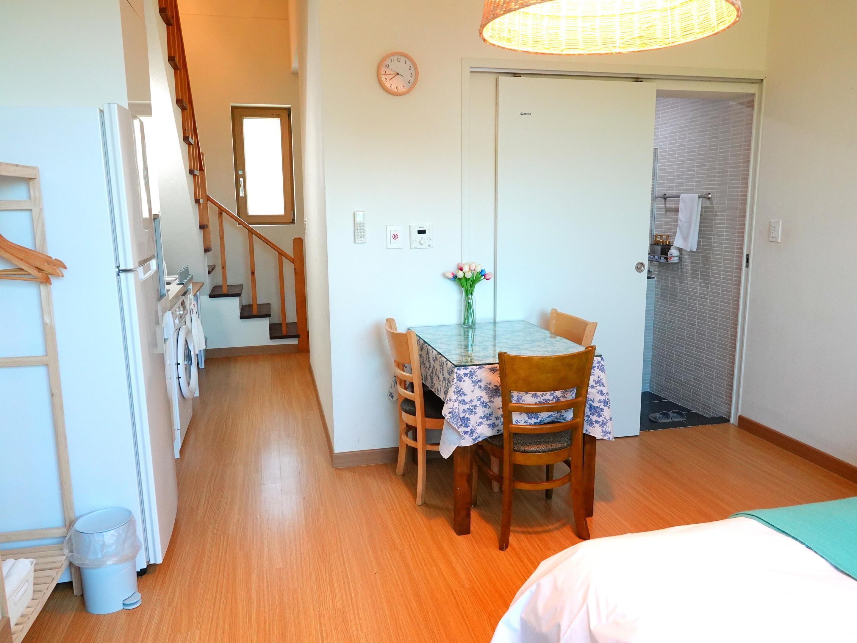 A compact kitchen with a wooden staircase, a dining area with a table and chairs, and a small bathroom with a towel rack.