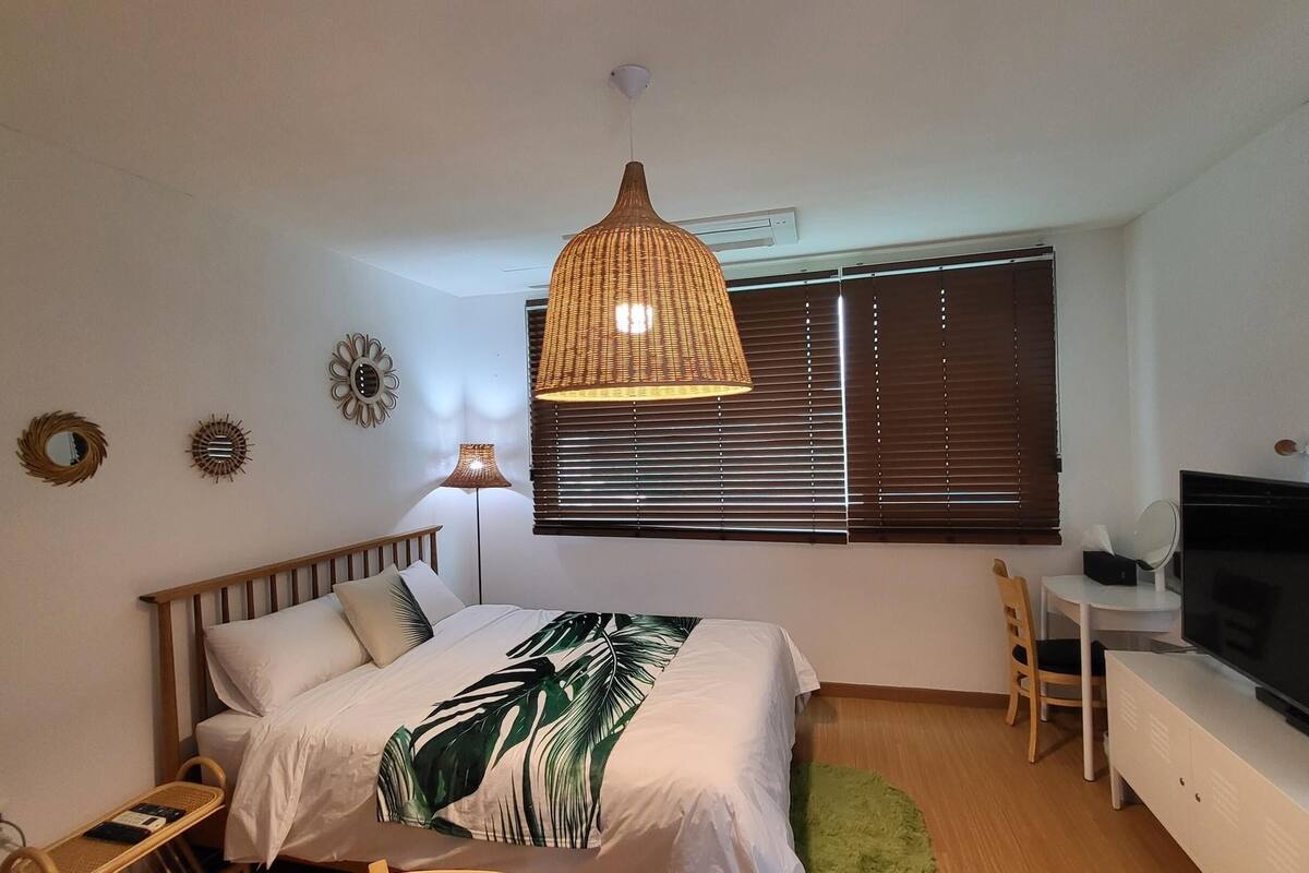 A bedroom with a bed, a TV, a desk, and a window with blinds.