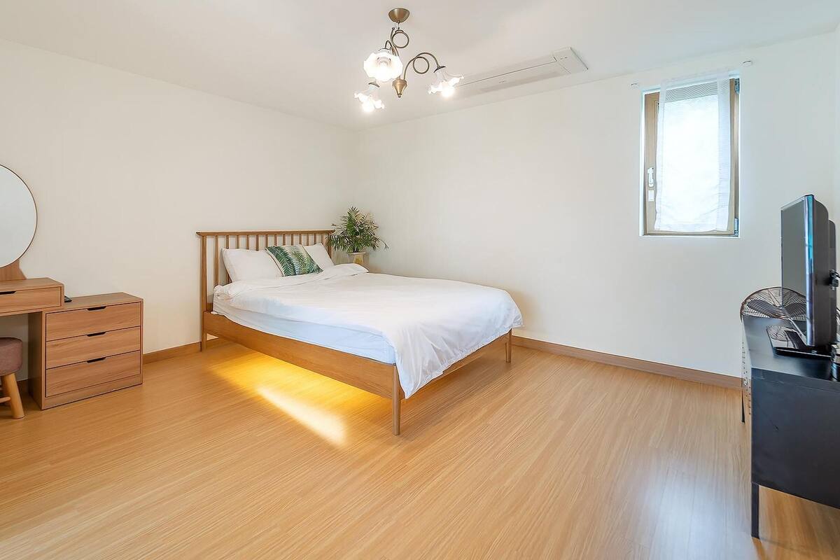 A bedroom with a wooden bed, a nightstand with a mirror, a dresser, a potted plant, and a TV.