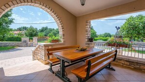 An outdoor patio with a wooden table and benches, stone pillars, and a view of greenery and buildings in the background. - Vacation home with pool - a paradise for families (Žminj)