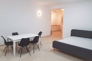A compact room with a white table and four black chairs, a single bed, and a kitchenette in the background.