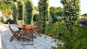 Eine Terrasse im Freien mit einem runden Tisch und vier Stühlen, umgeben von einem Garten mit blühenden Pflanzen und einer Kletterpflanzen-Gitterstruktur.
