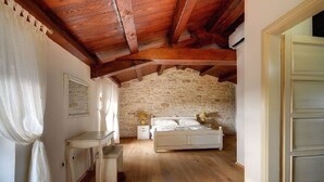 Een slaapkamer met een stenen muur, houten balken in het plafond, een bed, nachtkastjes en een witte deur.