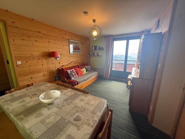 A cozy room with a wooden wall, a sofa, a dining table with a bowl, and a view through a window.