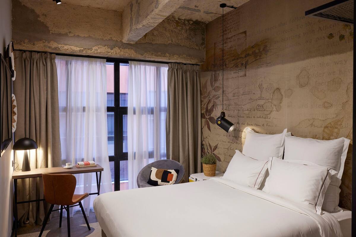 Een hotelkamer met een groot bed, een bureau met een lamp, een stoel en uitzicht door transparante gordijnen.
