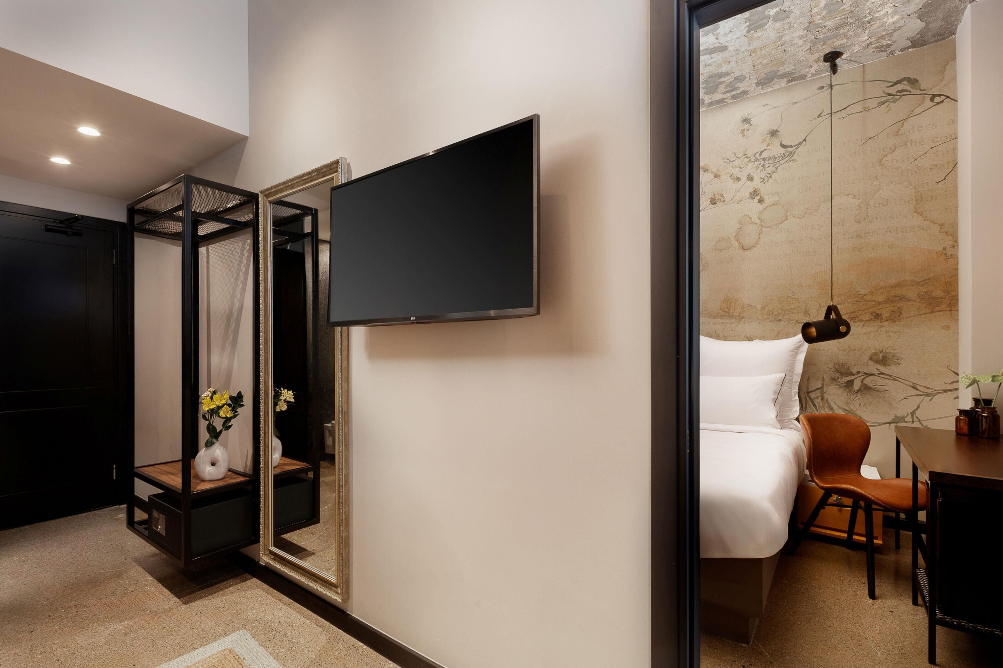 Une chambre d’hôtel moderne dotée d’un grand téléviseur à écran plat fixé au mur, d’un lit avec du linge de maison blanc, d’une table de chevet en bois, d’une petite table ornée d’un vase de fleurs et d’une chaise.