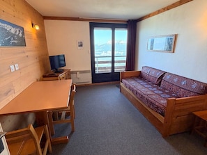 A room with a wooden table, a sofa, a television, and a view through a large window.
