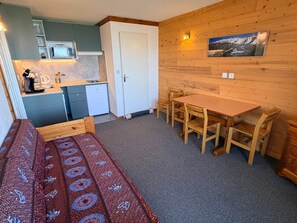 A compact room with a bed, a small kitchenette, a dining table with chairs, and a wooden wall with a picture.