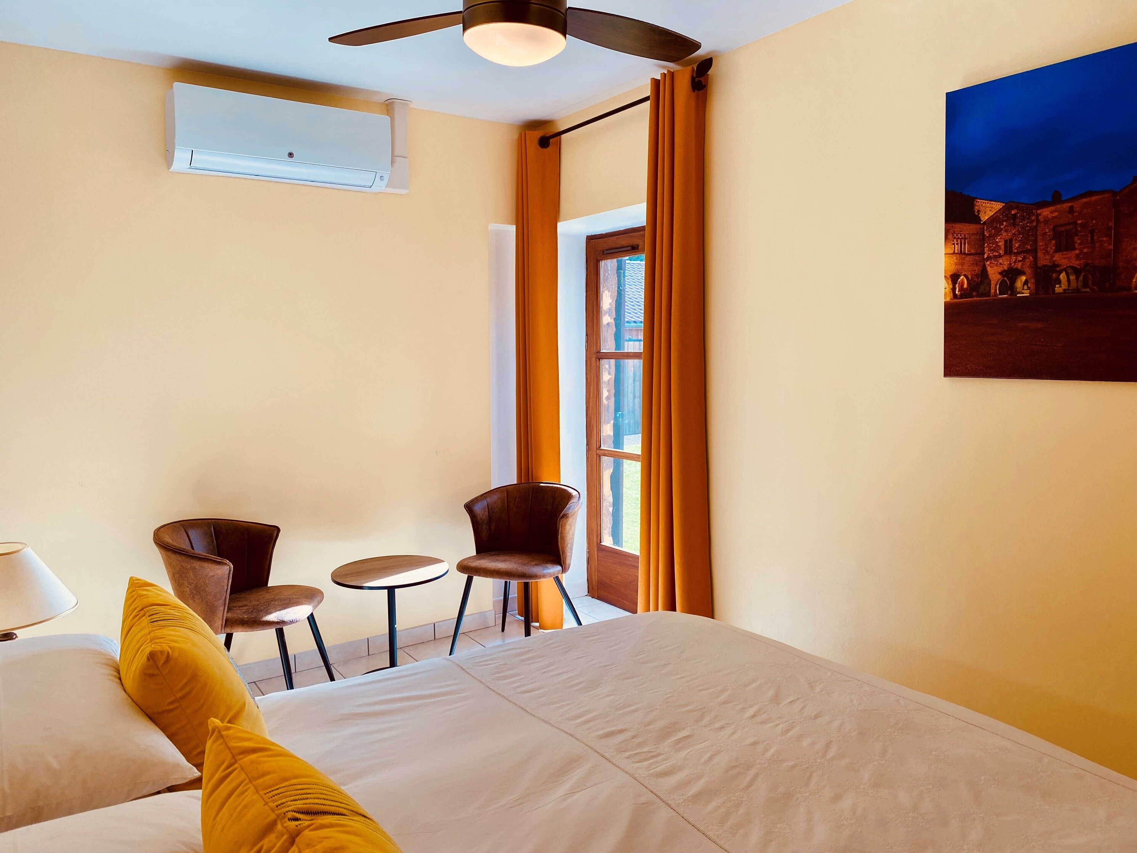 Een hotelkamer met een bed, twee stoelen, een tafel, een plafondventilator en een raam met gordijnen.