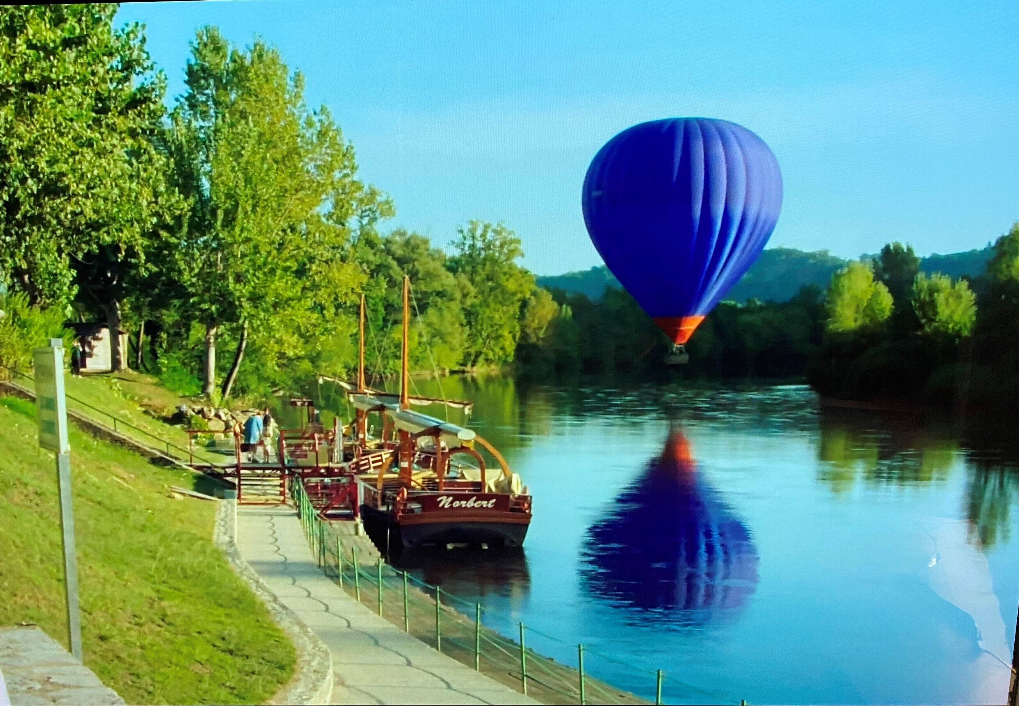 Eine Heißluftballon über einem ruhigen Fluss, ein Steg mit einem Boot und eine grüne Wiese mit Bäumen.