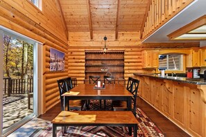 A log cabin interior with a dining area, kitchen, and a large window overlooking a forest.