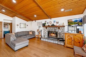 Un salon avec une cheminée en pierre, un canapé d’angle gris, un plancher en bois et un téléviseur.