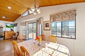 A dining area with a round table, chairs, a chandelier, and a view through large windows.