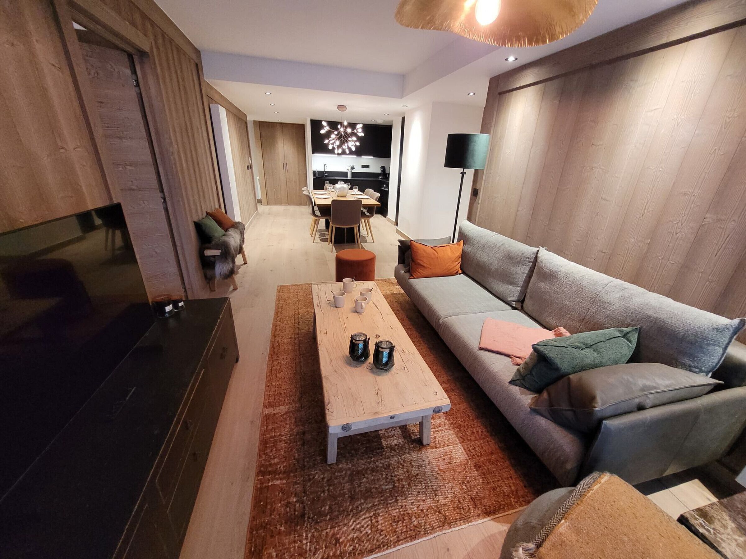 Una sala de estar moderna con un sofá, una mesa de centro y un televisor. Se aprecia un comedor al fondo.