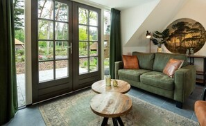 A living room with a green sofa, two round wooden tables, and a large window with a view of greenery. - Sprielderbosch 10 in ruhiger Lage in einem Luxuspark in der Nähe von Garderen (Putten)