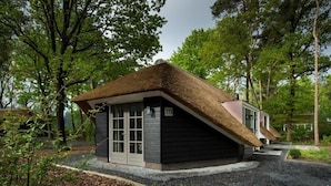 A small, dark-colored building with a thatched roof, surrounded by trees. - Sprielderbosch 10 quietly located in a luxury park near Garderen (Putten)