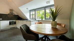 A modern kitchen with white cabinets, a black countertop, and a built-in oven. There's a round wooden table with chairs and a potted plant. - Sprielderbosch 10 quietly located in a luxury park near Garderen (Putten)