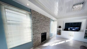 A living room with a stone fireplace, a flat-screen TV mounted on the wall, and a large window with blinds. - SERENITY @  ITS HEIGHTS (Cape Town)
