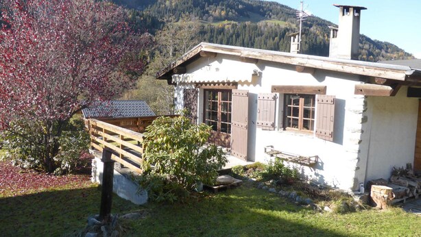 A rustic chalet with a wooden deck, a stone chimney, and a view of a forested mountain. - Unique chalet for 8 ppl, pets welcome, parking (Les Contamines-Montjoie)