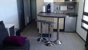 Eine kompakte Küche mit Mikrowelle, Toaster und Kaffeemaschine. Ein Barhocker und ein kleiner Esstisch sind vorhanden. Ein graues Sofa mit einem violetten Kissen befindet sich im Vordergrund.
