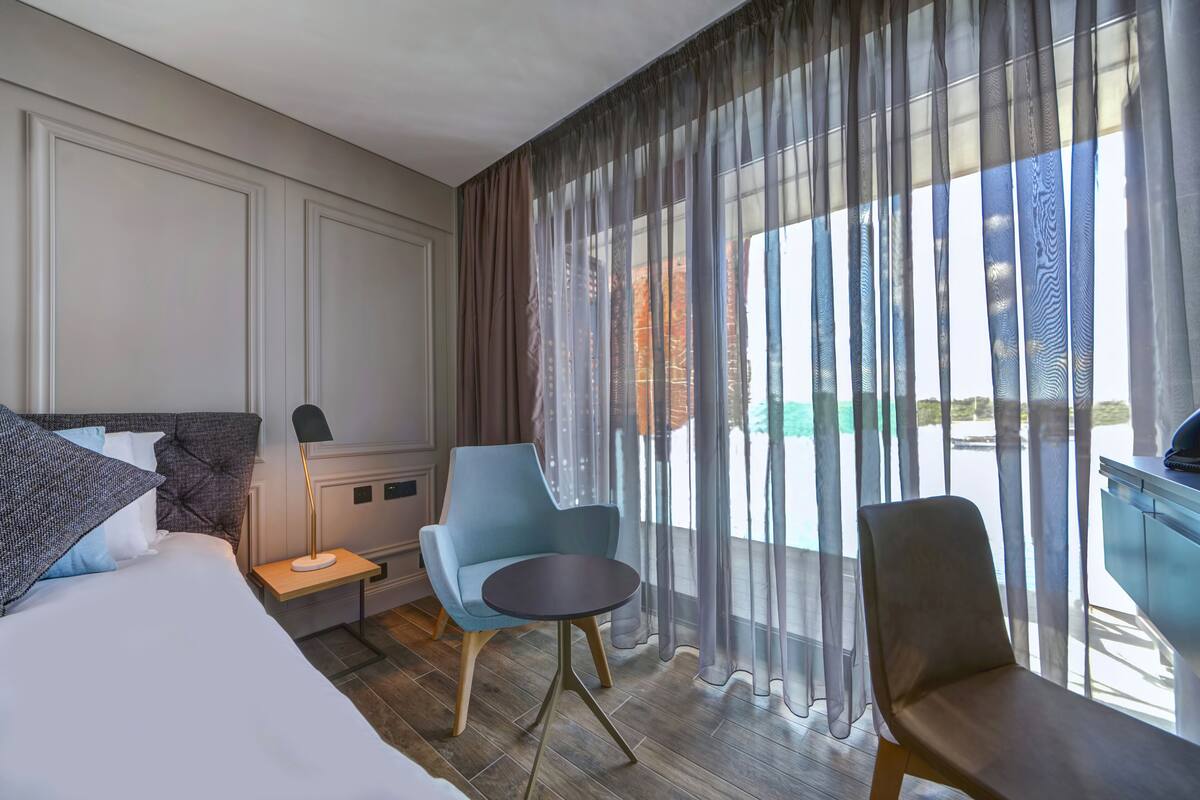 A hotel room with a bed, two chairs, a small table, and a view of a building through sheer curtains.