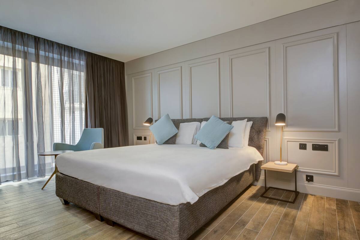 A modern hotel room with a large bed, a nightstand, a chair, and a view of the city through sheer curtains.