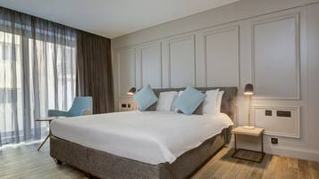 A modern hotel room with a large bed, a nightstand, a chair, and a view of the city through sheer curtains.