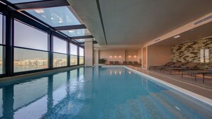 An indoor swimming pool with clear glass walls, lounge chairs, and a city view. - Land's End Boutique Hotel (Sliema)