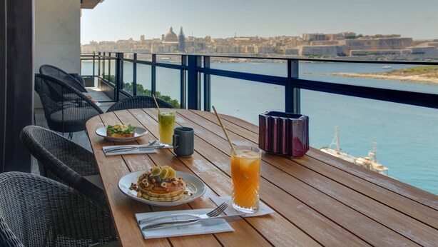 A wooden table with breakfast items and drinks, overlooking a waterfront cityscape. - Land's End Boutique Hotel (Sliema)