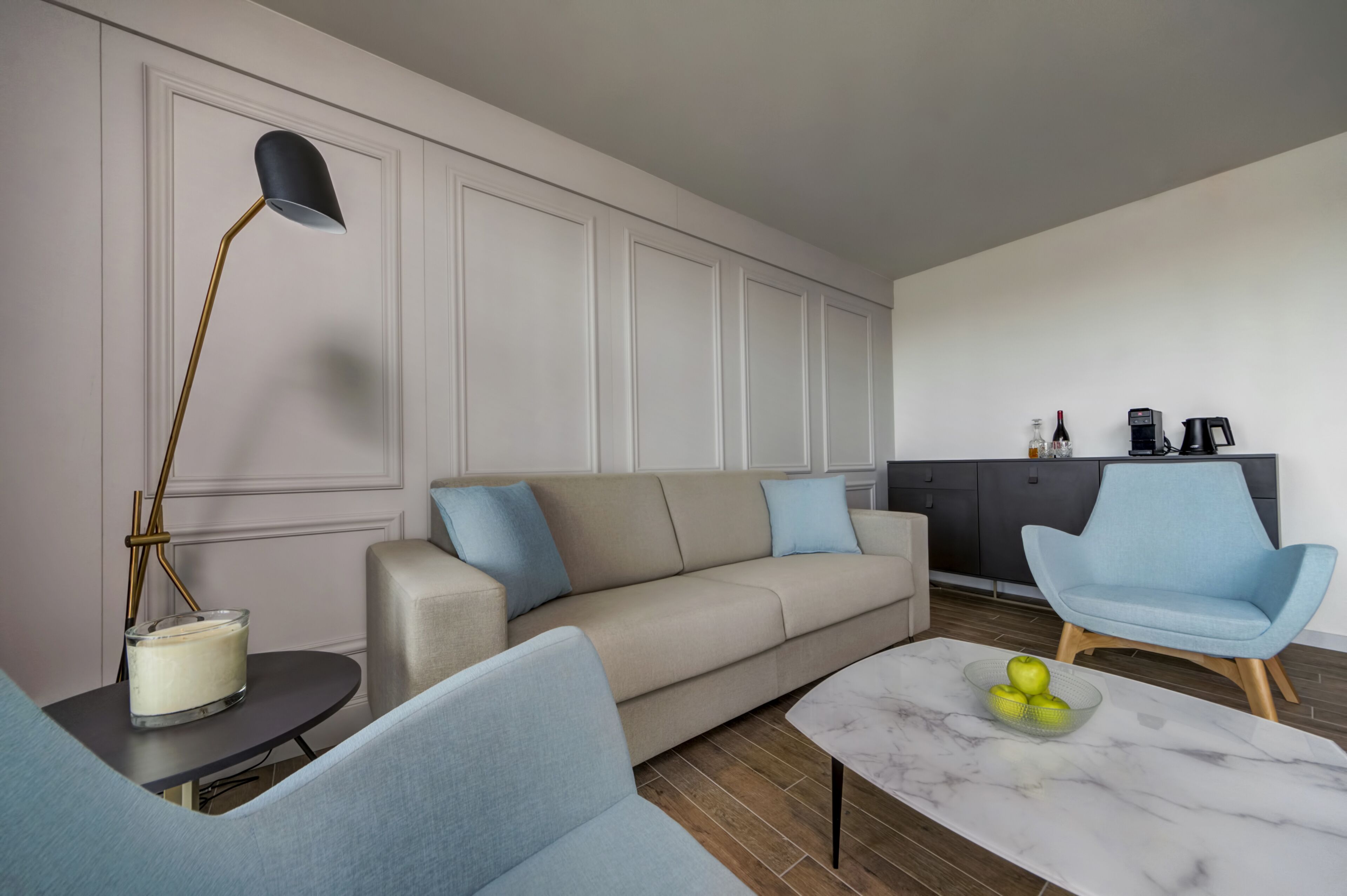 Un salón moderno con un sofá color beige, sillones azules, una mesa de centro de mármol con manzanas, una lámpara de pie y una zona de cocina con un mueble negro y una cafetera.