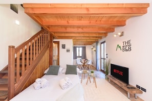 Una camera d'albergo con una scala in legno, un letto con asciugamani arrotolati, un tavolino con una pianta, una TV su un mobile e un cartello a parete con la scritta "THE MOUNTAIN B&B".