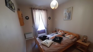 Een slaapkamer met een houten bed, een nachtkastje, een hanglamp, een raam met gordijnen en een aan de muur hangend schilderij.