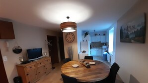 Een moderne keuken met witte kasten, een eethoek met een houten tafel en zwarte stoelen, en een grote klok aan de muur.