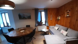 Een gezellige woonkamer met een houten wand, een ronde eettafel met stoelen, een grijze bank met kussens en een schilderij aan de muur.