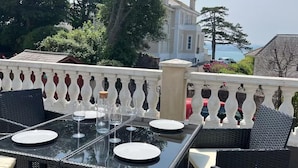 Un área de comedor al aire libre con una mesa preparada para dos, rodeada de sillas de mimbre, una barandilla y vistas a un edificio y árboles.