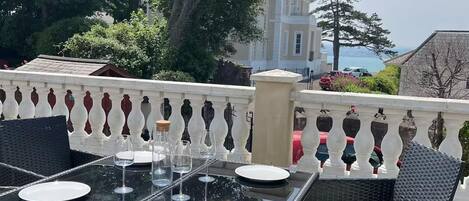 An outdoor dining area with a table set for two, surrounded by wicker chairs, a balustrade, and a view of a building and trees.