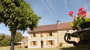 Ein zweistöckiges Steinhaus mit Ziegeldach, ein großer Baum im Vordergrund und eine Topfpflanze mit rosa Blüten im Hintergrund.