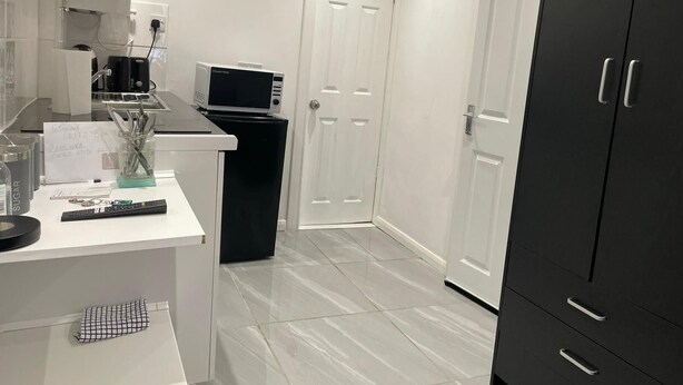 A modern kitchen with white cabinetry, a black refrigerator, a microwave, and a coffee machine. There is a door with a handle and a small clock on the wall. - Onsite Cosy Practical Studio - in a Quite Area (Coventry)