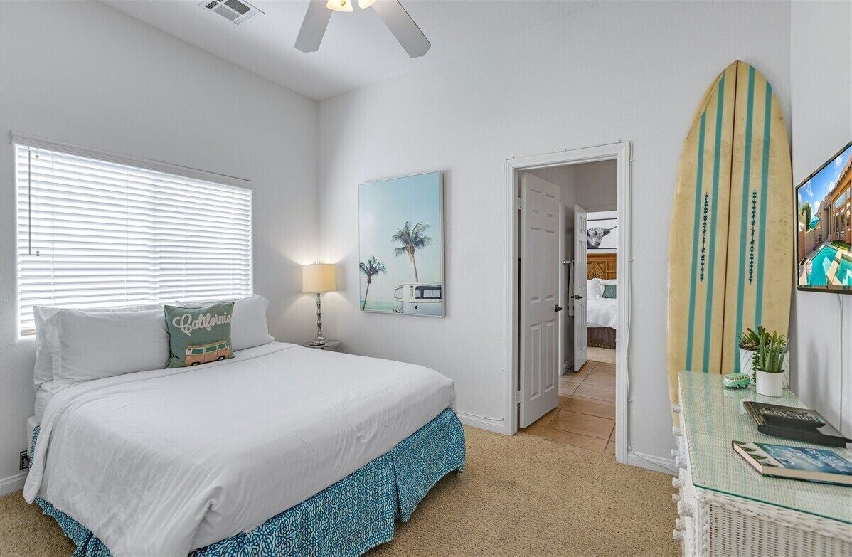 A bedroom with a bed, a nightstand, a lamp, a framed picture, a surfboard, and a door leading to another room.