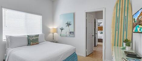 A bedroom with a bed, a nightstand, a lamp, a framed picture, a surfboard, and a door leading to another room.
