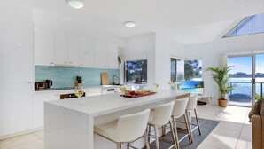 Een moderne keuken met een centraal eiland, barstoelen en uitzicht op zee door grote ramen.