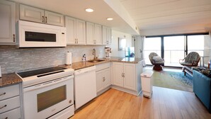 A modern kitchen with white cabinetry, a built-in microwave, and a dishwasher. There is a large window providing natural light and a view of the outdoors. - Beautiful 2-bedroom oceanfront condo with gorgeous ocean views, free WiFi, and an outdoor pool located uptown with direct access to the beach! (Ocean City)
