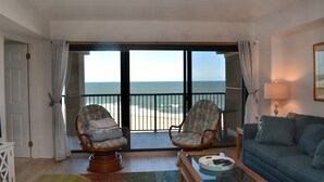 A living room with a view of the ocean, two wicker chairs, a glass coffee table, and a blue sofa. - Beautiful 2-bedroom oceanfront condo with gorgeous ocean views, free WiFi, and an outdoor pool located uptown with direct access to the beach! (Ocean City)