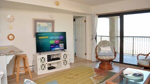 A living room with a flat-screen TV, a wicker chair, a dining table, and a view of the outside through a sliding glass door. - Beautiful 2-bedroom oceanfront condo with gorgeous ocean views, free WiFi, and an outdoor pool located uptown with direct access to the beach! (Ocean City)