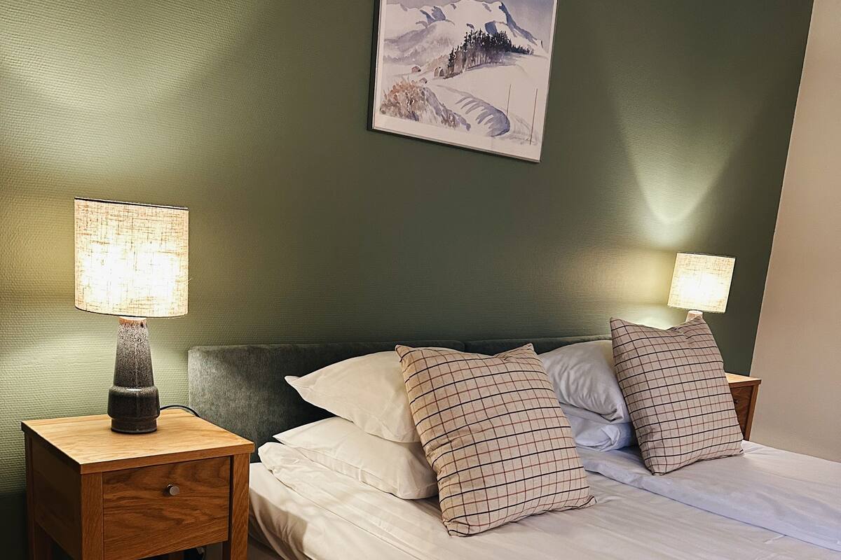 A neatly made bed with white linens and two pillows, a wooden nightstand with a lamp, and a painting of a snowy mountain landscape on the wall.