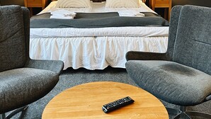 Habitación de hotel con cama, dos mesitas de noche con lámparas, una mesa redonda de madera y un mando a distancia, y dos sillones grises.
