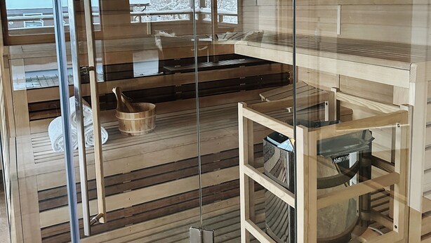 Una habitación moderna con paredes de vidrio, suelo de madera y mobiliario, incluyendo un banco y una pequeña mesa con un recipiente.